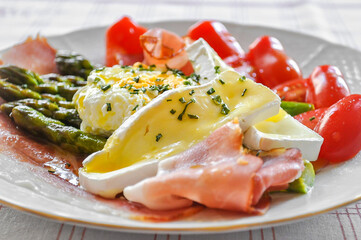 Delicious green boiled asparagus, ham, prosciutto, grape tomatoes, poached organic egg and brie cheese all seasoning with herbs on the white plate. Light tasty breakfast, brunch or dinner.