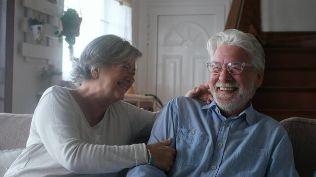 Portrait of couple of seniors laughing and having fun together sitting on the sofa of home indoor. Two old and mature people smiling and enjoying joking.
