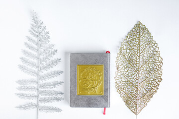 Flat lay gold Arabic on book of holy Quran between silver and gold leaves on white background