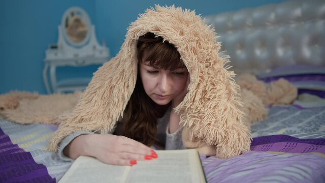 brunette woman lies on the bed and reads a book, tited and covered with a blanket