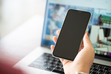Mockup image of woman hand using black mobile phone with laptop screen