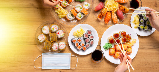 Banner. Lockdown home dinner party in pandemic with Japanese sushi from delivery or takeaway service. Various salmon, crab, vegan rolls, wasabi, ginger on wooden background, medical mask. Top view