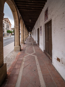 Perspective view looking through the historic stone archway arcades of the Plaza de la Duquesa de Parcent in Ronda, Spain, with a paved tile walkway and rustic wooden doors.