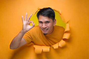 Young entertaining man through torn yellow paper hole showing okay sign with facetious of content and happiness.