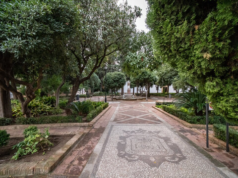 The peaceful, shaded gardens of Plaza de la Duquesa de Parcent in Ronda, Spain, featuring a decorative tiled pavement walkway, lush green trees, and a central fountain.