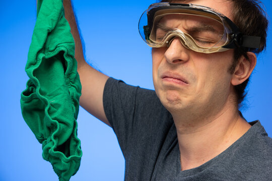 Caucasian male wearing goggles making disgusted face gesture at smelly green underwear held in hand studio shot