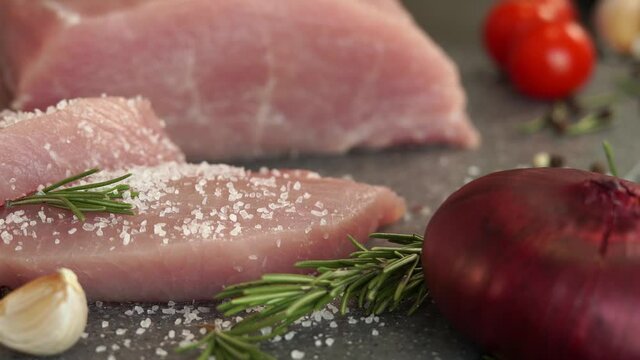 Close up of fresh pork is sprinkled with salt on background of onion, garlic, greens and spices. Food concept, cooking meat, loin, carbonate.