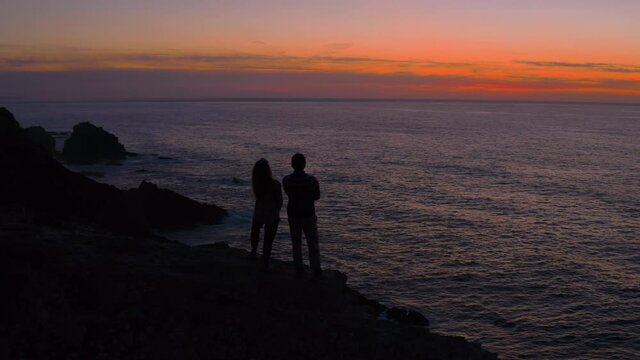 Aerial drone shot of silhouette of couple stand on edge of cliff during epic orange sunset. Inspiring and motivational adventure travel wanderlust video. Beautiful nature exploration and tranquility