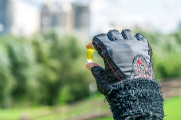 Close-up view of female hand in black glove holding yellow fish oil capsule. Blurred summer city...