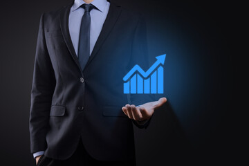 Businessman holding tablet and showing a growing virtual hologram of statistics, graph and chart with arrow up on dark background. Stock market. Business growth, planing and strategy concept.