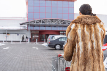 A woman in a fur coat goes to a large supermarket with a shopping basket. Winter time of the year. - obrazy, fototapety, plakaty