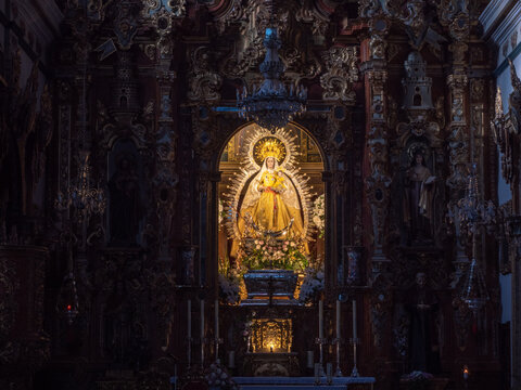 Interior view of a dimly lit church sanctuary highlighting a brilliant, illuminated golden altarpiece retable centered on a statue of the Virgin Mary with a radiant starburst halo, housed within an in