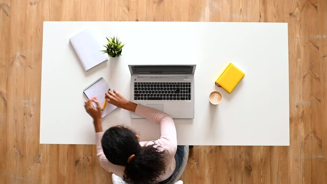 Overhead view of mixed-race ethnic woman or female freelancer with curly hair sitting at the white table desk with well-organized space, typing on the laptop, taking notes, modern home office concept