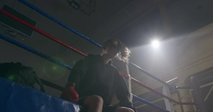 Tired female boxer sitting on boxing ring after kickboxing workout in the gym. Exhausted woman athlete resting on ring ropes. Low angle shot.