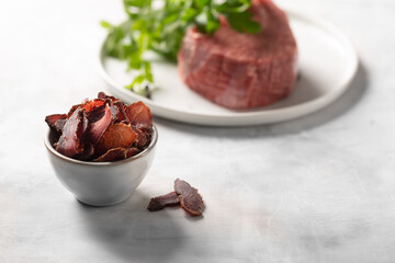 Bowl with jerky meat and raw beef on gray concrete background