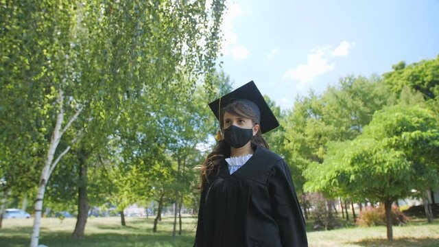 A girl student graduate in a graduation gown in a protective medical mask walks alone in the park during the coronavirus pandemic.