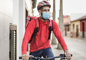 Naklejka premium Young caucasian man working for food delivery with electric bicycle while wearing safety mask for coronavirus outbreak