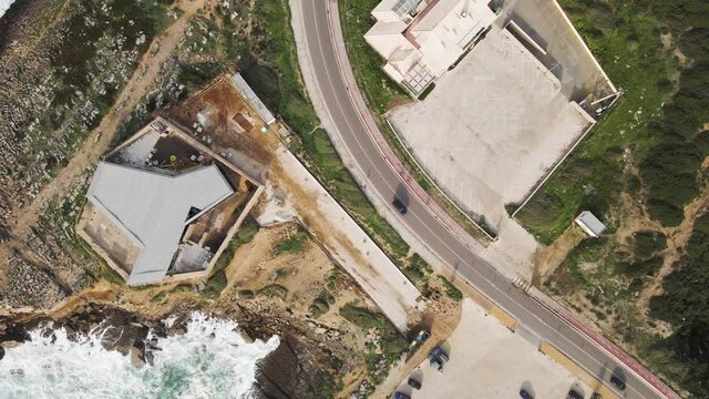 Aerial view of a desertic road driving next to a view of rough sea with crispy waves. Place situated in the south Portuguese wild coastline between the Atlantic ocean and the forest. Cascais, Portugal