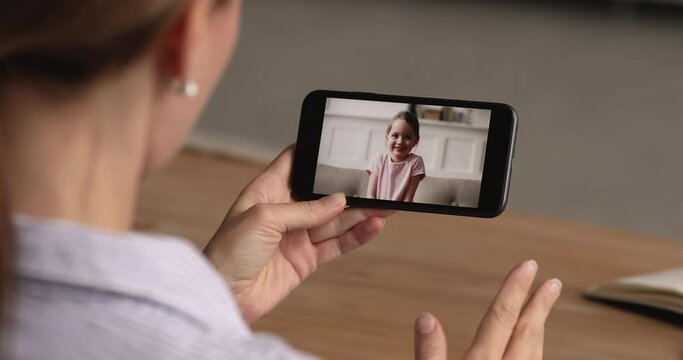 Shoulder close up view young woman holding telephone in hands, holding video call web camera zoom conversation with adorable little 7s kid daughter, sharing life news, enjoying distant communication.