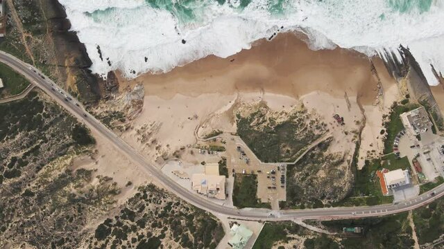 Aerial view of a desertic road driving next to a view of rough sea with crispy waves. Place situated in the south Portuguese wild coastline between the Atlantic ocean and the forest. Cascais, Portugal