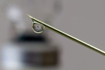 Drop of vaccine against Covid-19 (SARS-COV2) coming out of a syringe. A vial of vaccine is reflected in the drop.