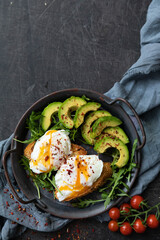 Breakfast with poached egg, avocado and arugula. with fried toast and cherry tomatoes, served in a cast-iron pan on a textile background. Top view copy space