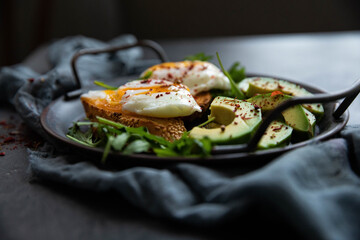 Delicious and delicious breakfast with poached egg on a slice of fried bread, avocado, arugula, cherry tomatoes and spices. Served in a cast-iron skillet, served with textiles on a black background . 