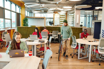 Bearded office manager holding bicycle while moving between desks of colleagues
