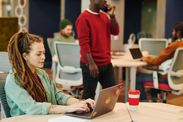 Young Caucasian businesswoman with dreadlocks sitting by desk in front of laptop