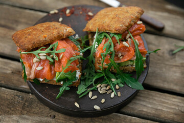 A delicious snack for lunch. Delicious sandwich with lightly salted salmon. With a lush bun, with arugula greens and sunflower seeds! Served on a wooden platter and rustic background. Horizontal orien