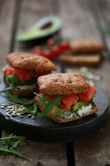 A delicious snack for lunch. Delicious sandwich with lightly salted salmon. With a lush bun, with arugula greens and sunflower seeds! Served on a wooden platter and rustic background. Horizontal orien
