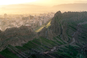 City of Qom, Iran, in a dramatic sunset viewed from the hill nearby. Steep rock cliffs in the front. Desert rocks, dramatic terrain.