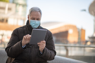 Covid coronavirus lifestyle, masked elder man using his tablet while sitting outdoor in a city square