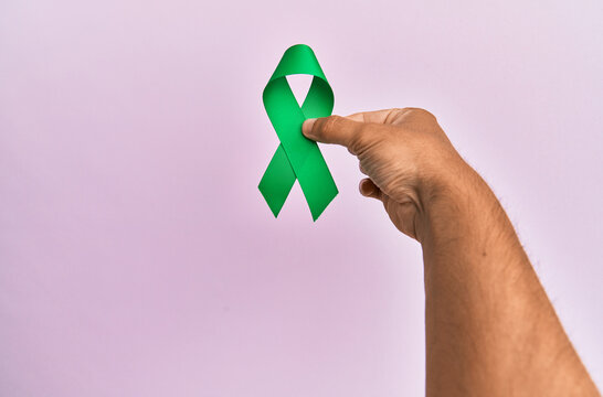 Hand of young hispanic man holding green ribbon over isolated pink background.