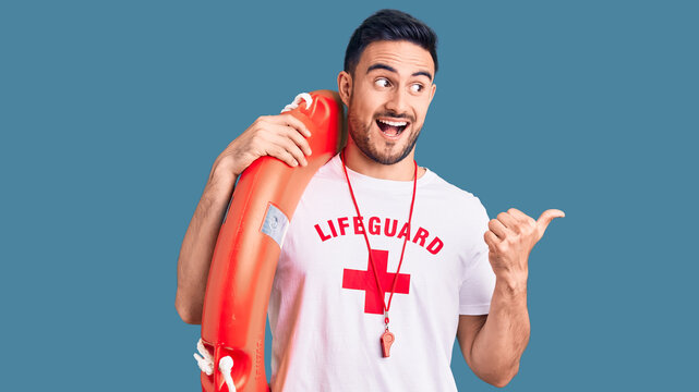 Young handsome man wearing lifeguard uniform holding float pointing thumb up to the side smiling happy with open mouth
