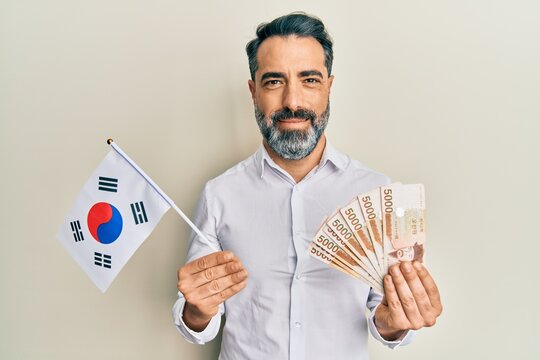 Middle Age Man With Beard And Grey Hair Holding South Korea Flag And Won Banknotes Smiling With A Happy And Cool Smile On Face. Showing Teeth.