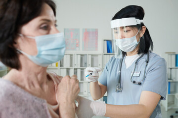 Nurse of Asian ethnicity in protective workwear making injection to mature woman