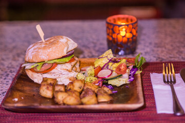 A fish burger on a wooden plate with sauteed potatoes and green salad. the meal is on a candle lit table