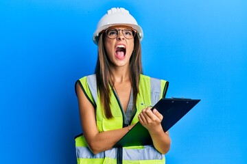 Young hispanic woman wearing safety helmet holding clipboard angry and mad screaming frustrated and furious, shouting with anger looking up.