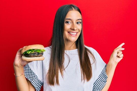 Young hispanic woman eating hamburger smiling happy pointing with hand and finger to the side