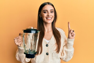 Beautiful brunette young woman holding food processor mixer machine smiling with an idea or question pointing finger with happy face, number one