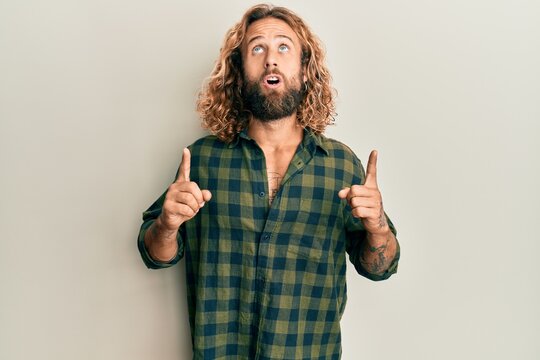 Handsome man with beard and long hair wearing casual clothes amazed and surprised looking up and pointing with fingers and raised arms.