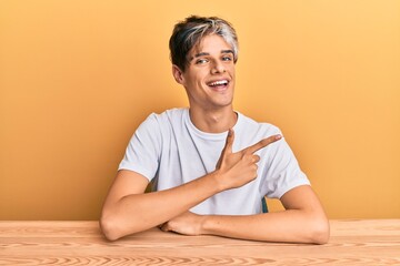 Young hispanic man wearing casual clothes sitting on the table smiling cheerful pointing with hand and finger up to the side