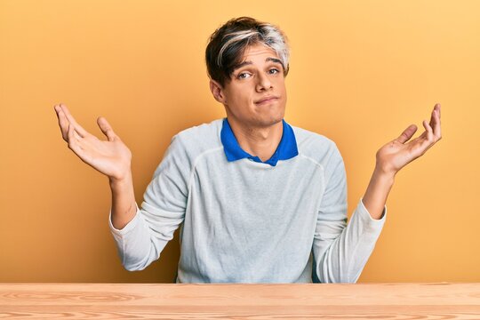 Young hispanic man wearing casual clothes sitting on the table clueless and confused with open arms, no idea and doubtful face.