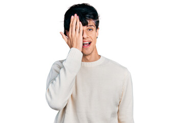 Handsome hipster young man wearing casual winter sweater covering one eye with hand, confident smile on face and surprise emotion.