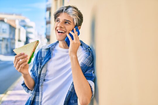 Young hispanic man smiling happy talking on the smartphone and eating sandwich at the city.