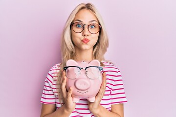 Beautiful caucasian blonde girl holding piggy bank with glasses puffing cheeks with funny face....