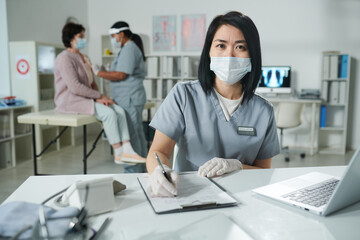 Fototapeta na wymiar Young Asian clinician in protective workwear sitting by desk and filling in document