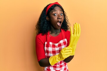 Beautiful african young woman wearing cleaner apron and gloves afraid and shocked with surprise and amazed expression, fear and excited face.