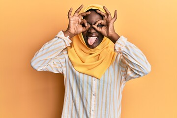 Beautiful african young woman wearing traditional islamic hijab scarf doing ok gesture like binoculars sticking tongue out, eyes looking through fingers. crazy expression.